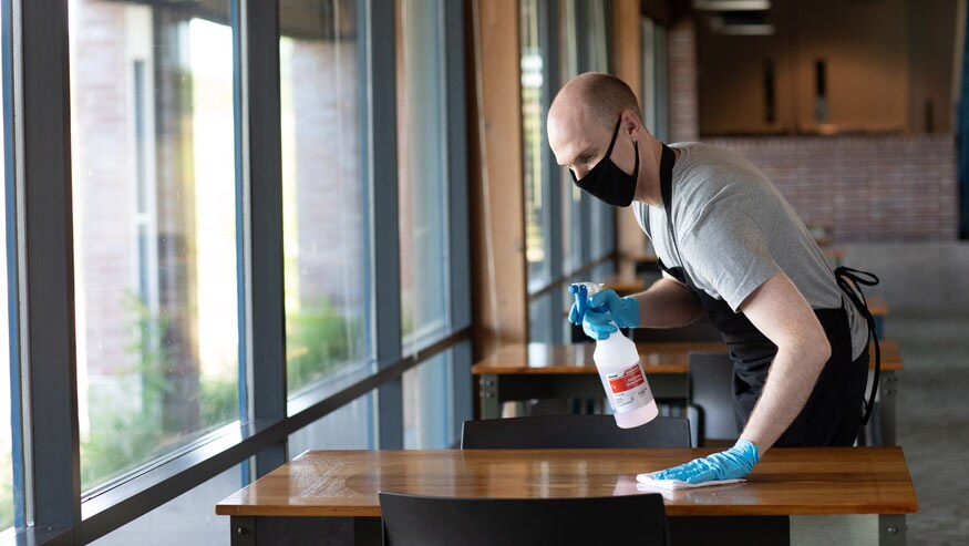 A glove-wearing waiter or busser wipes down a restaurant table with a spray bottle of Sink and Surface Cleaner Sanitizer, a 2-in-1 food contact surface cleaner sanitizer concentrate by Ecolab.